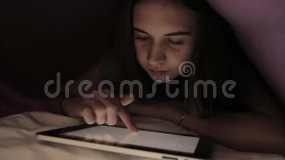 年轻女孩躲在羽绒被下在黑暗中使用PC平板设备青少年晚上在数字平板电脑上玩视频的预览图