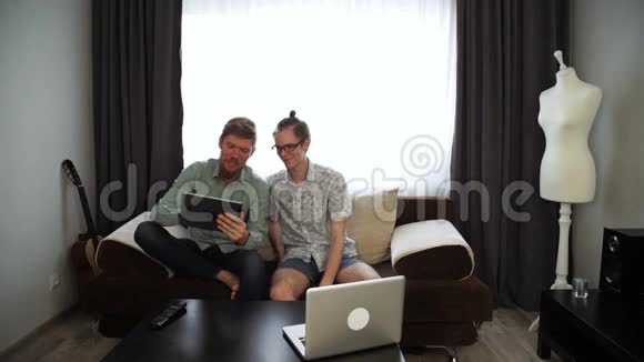 两个年轻人的兄弟正在家里工作坐在沙发上在他的平板电脑上在台式笔记本电脑上工作视频的预览图