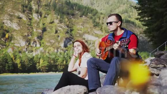 一对在海边玩吉他的情侣在山河旁边的一个3840x2160视频的预览图