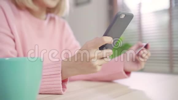 美丽的亚洲女人使用智能手机通过信用卡购买网上购物而穿毛衣坐在客厅的桌子上视频的预览图
