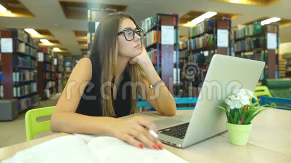 在图书馆里无聊的学生在图书馆里的电脑上工作带眼镜的深色头发视频的预览图