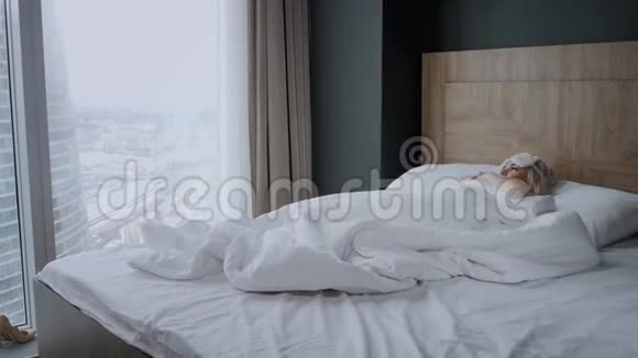 早上在酒店房间年轻女子坐在舒适的床上戴着面具睡在头上摩天大楼的窗户视频的预览图