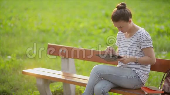 可爱的年轻女人在公园里拿着平板电脑漂亮的女人在花园里的长凳上拿着平板电脑年轻的女学生视频的预览图