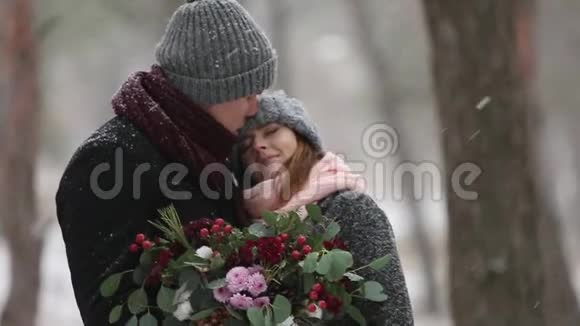新婚夫妇新郎和新娘拥抱亲吻并在降雪期间在雪松林中温暖对方青年视频的预览图