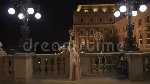 美丽的女士穿着一件优雅的粉红色长裙晚上在老大理石城堡的阳台上摆姿势视频的预览图