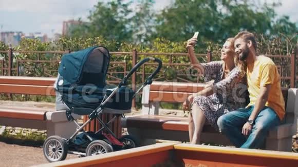 年轻的父母带着婴儿车坐在长凳上在智能手机上自拍家庭视频的预览图