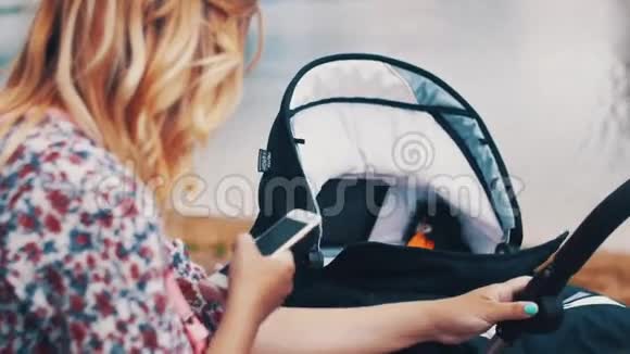母亲摇摆婴儿车和使用智能手机在湖边夏日阳光明媚视频的预览图