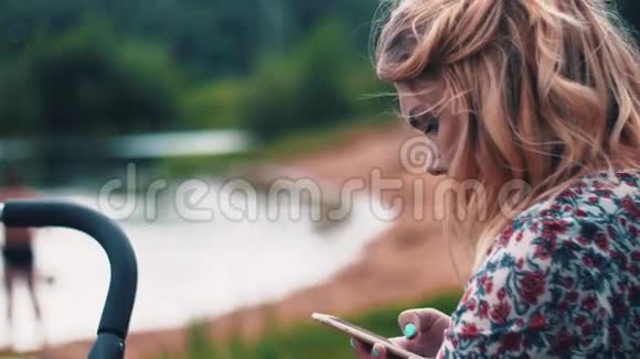 母亲摇摆婴儿车和使用智能手机在湖边夏日步行视频的预览图