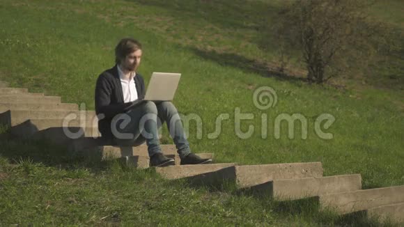 利用笔记本电脑坐在公园楼梯上的年轻人的侧景视频的预览图