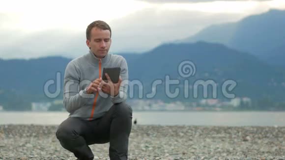 年轻英俊的男人坐在岩石海滩上他检查平板电脑上的信息在太平洋和视频的预览图