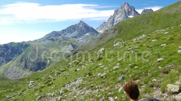 女人在田园诗般的山景中跋涉在小径上穿越盛开的绿色草地在高高的岩石山中响起视频的预览图