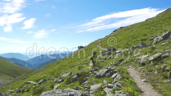 女人在田园诗般的山景中跋涉在小径上穿越盛开的绿色草地在高高的岩石山中响起视频的预览图