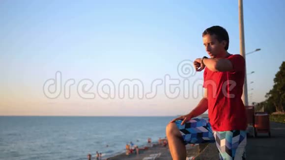 年轻英俊的男人带着智能手表在海边放松他在日落时分查看社交网络的信息视频的预览图