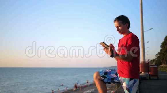 年轻的帅哥在海边用平板电脑放松他在日落时分查看社交网络上的信息视频的预览图