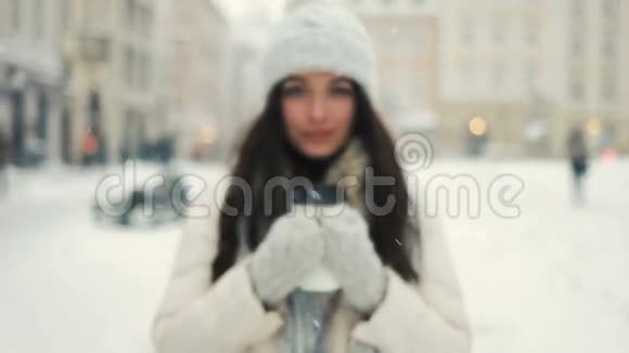 微笑的年轻女子穿着白色温暖的衣服喝着咖啡带走白雪覆盖的城市背景视频的预览图