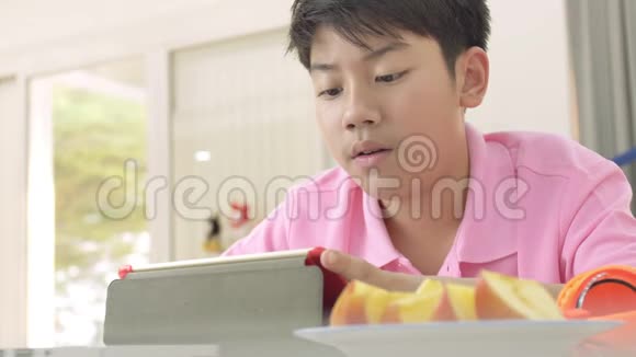 快乐的亚洲男孩玩平板电脑男孩吃一块苹果看平板电脑视频的预览图