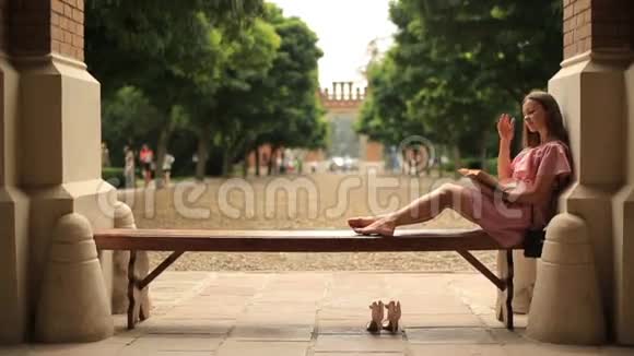 在大学校园里一个年轻漂亮的女孩躺在长凳上赤脚看书视频的预览图