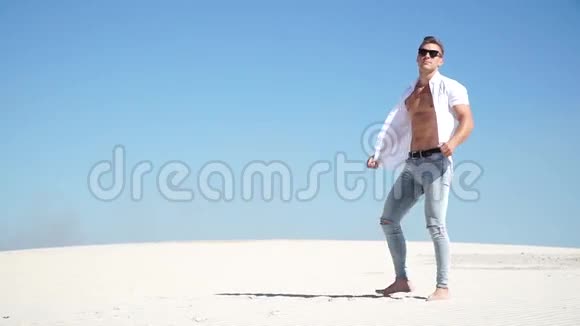 穿牛仔裤的时髦男人戴着眼镜穿着白衬衫光着脚站在沙漠里的沙滩上视频的预览图