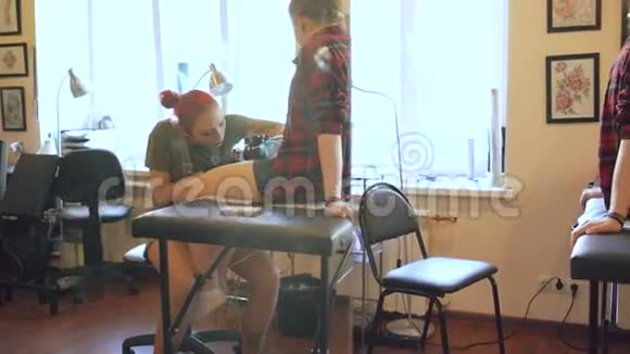 美丽的红发女子纹身艺术家纹身图片在年轻女孩客户的腿在工作室视频的预览图