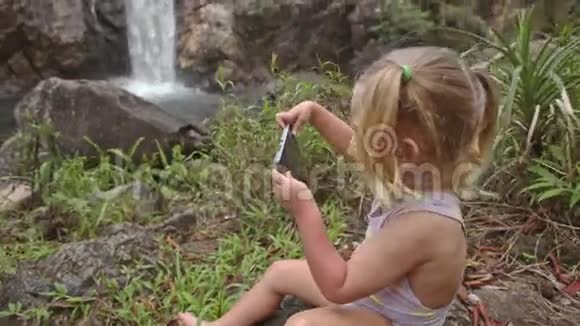 特写小女孩在石头上打电话对抗瀑布视频的预览图
