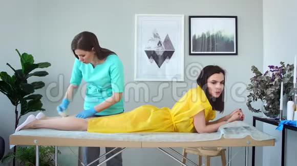 两个女孩在一家美容院主人穿着浴衣戴着手套做着用糖去除腿上毛发的程序视频的预览图