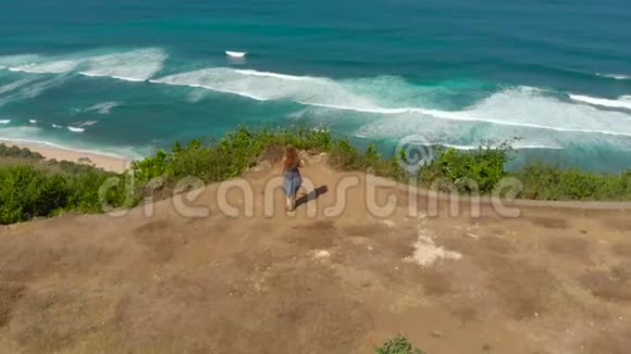 一名年轻女子在巴厘岛的一个偏僻的海滩上游览的空中镜头站在岩石上看着一个视频的预览图