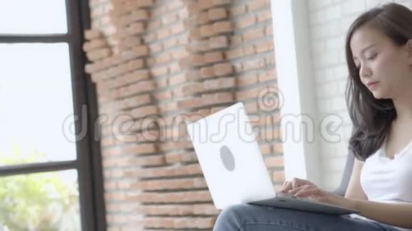 美丽的肖像亚洲年轻女子微笑着在网上笔记本电脑工作快乐地坐在客厅的沙发上视频的预览图