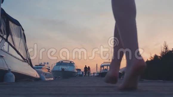 日落时分穿着蓝色连衣裙的年轻女孩在海岸的木墩上散步白色游艇视频的预览图