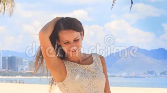 穿着白色连衣裙的苗条女孩微笑着抚平海滩上的头发视频的预览图