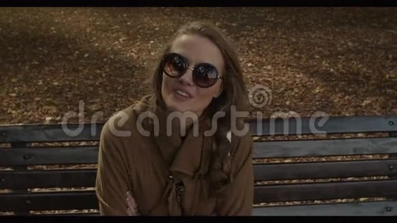 秋天公园里坐在长凳上戴着太阳镜的漂亮年轻女子视频的预览图