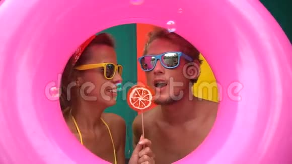有趣的年轻夫妇吃肥皂泡和舔橙色棒棒糖隔离在粉红色充气环中视频的预览图