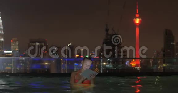 在摩天大楼屋顶上的游泳池中的女性景观使用平板电脑对抗夜间城市景观马来西亚吉隆坡视频的预览图