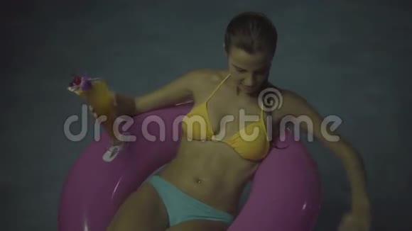 穿比基尼的快乐女人晚上在游泳池里喝鸡尾酒在充气环上游泳视频的预览图