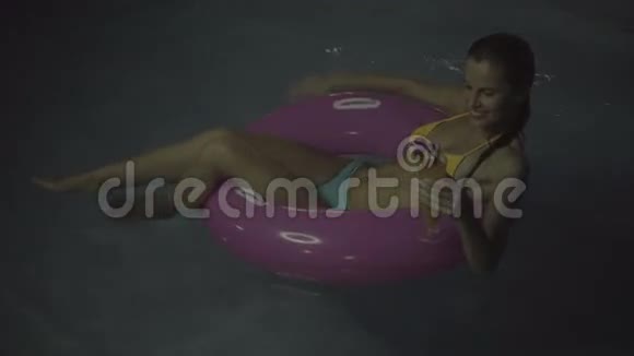 穿比基尼的快乐女人晚上在游泳池里喝鸡尾酒在充气环上游泳视频的预览图