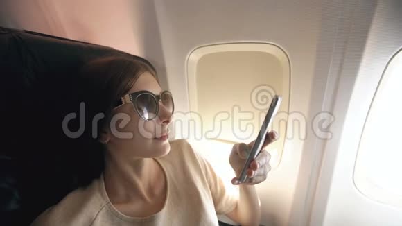 日落时分坐在飞机窗口附近的旅游妇女在飞行中使用手机视频的预览图