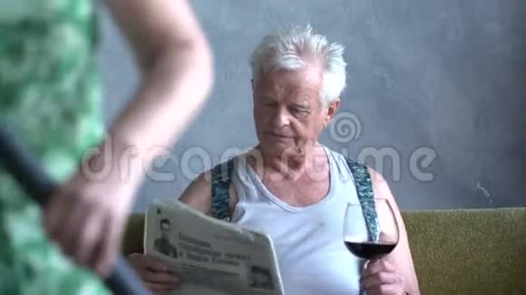 有趣的老人在家里喝酒在妻子的背景下用吸尘器看报纸视频的预览图