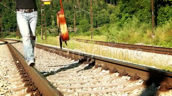 一个穿破牛仔裤的年轻吉他手沿铁轨行走的慢镜头视频的预览图