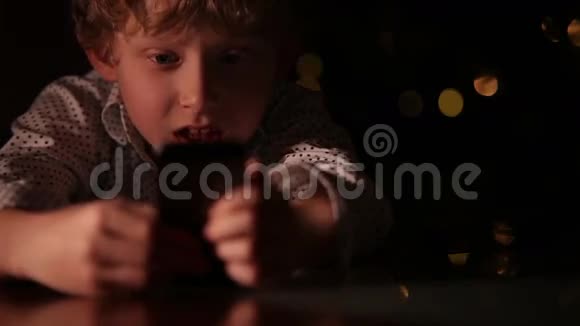 卷发小男孩玩智能手机背景是圣诞树灯视频的预览图