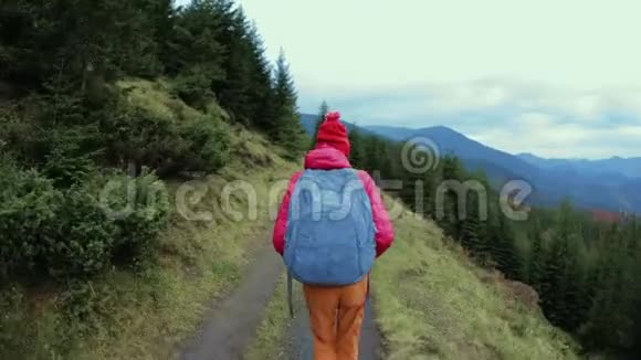 女徒步旅行者带着背包穿着红色夹克和橙色裤子走在山上的背景上视频的预览图