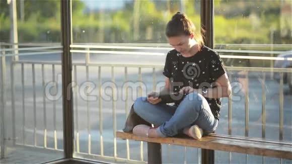 年轻迷人的白人女性穿着时髦的服装在公共汽车站等待交通时使用平板电脑女人坐在公共汽车站上视频的预览图