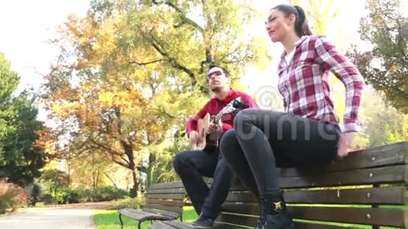 男人弹吉他女人跟着节奏跳舞坐在公园的长椅上视频的预览图