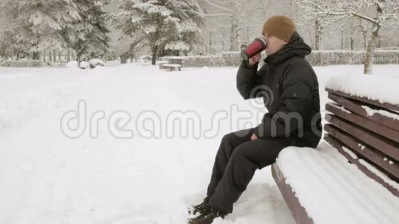 一个年轻人在冬天的公园里喝热水瓶里的热饮料大雪纷飞穿着深色夹克和保暖衣服的男人视频的预览图