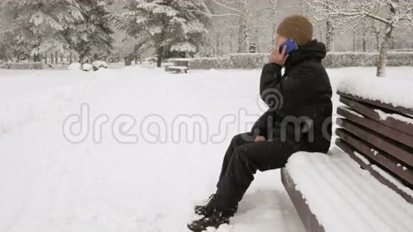 冬天公园的一个年轻人在打电话他钦佩雪和树的两边一个穿深色夹克的男人视频的预览图