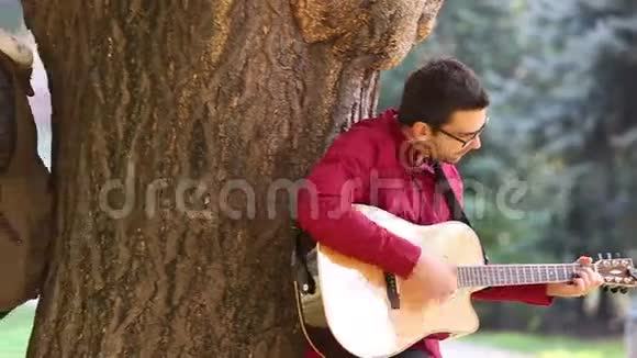 年轻夫妇一边弹吉他一边唱歌一边倚在公园的树上视频的预览图