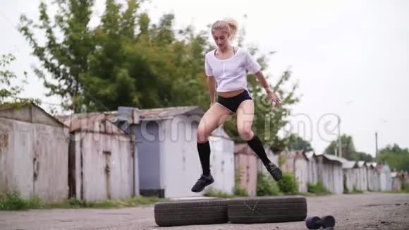 运动的年轻金发女子穿短裤在轮胎俯卧撑跳跃的帮助下进行各种力量练习视频的预览图