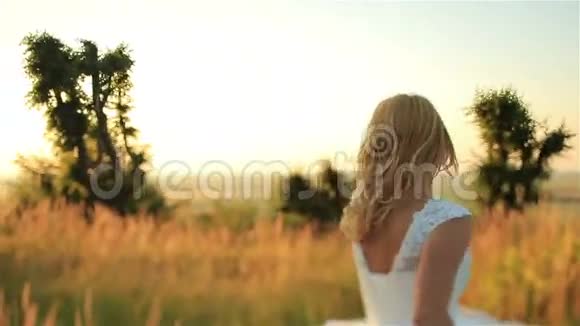 日落时分美丽的金发新娘穿着长长的新娘礼服穿过黑麦田视频的预览图