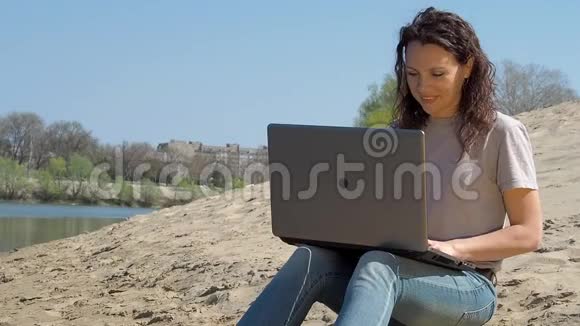 带着笔记本电脑的女孩在河边的新鲜空气中工作春天阳光明媚的一天视频的预览图