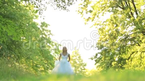 快乐浪漫时尚的金发新娘穿着白色连衣裙在阳光下在森林里奔跑和旋转视频的预览图