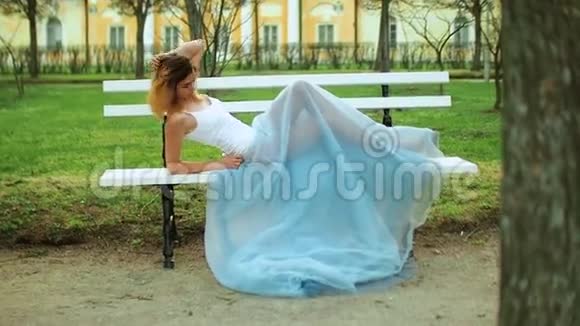 漂亮的女孩穿着白色和蓝色的连衣裙躺在公园路的长凳上在照片拍摄摆姿势视频的预览图