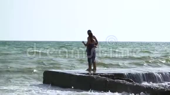 一对美丽的年轻夫妇在暴风雨中走在海边波浪在码头上破浪前进一个穿着泳裤的男人一个穿着泳裤的女孩视频的预览图